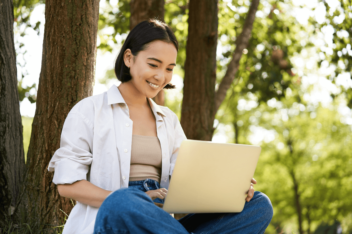 A woman happily works from her laptop while sitting on the ground in a park