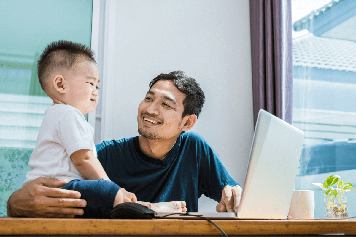 A dad takes a break from working on his laptop to smile at his toddler