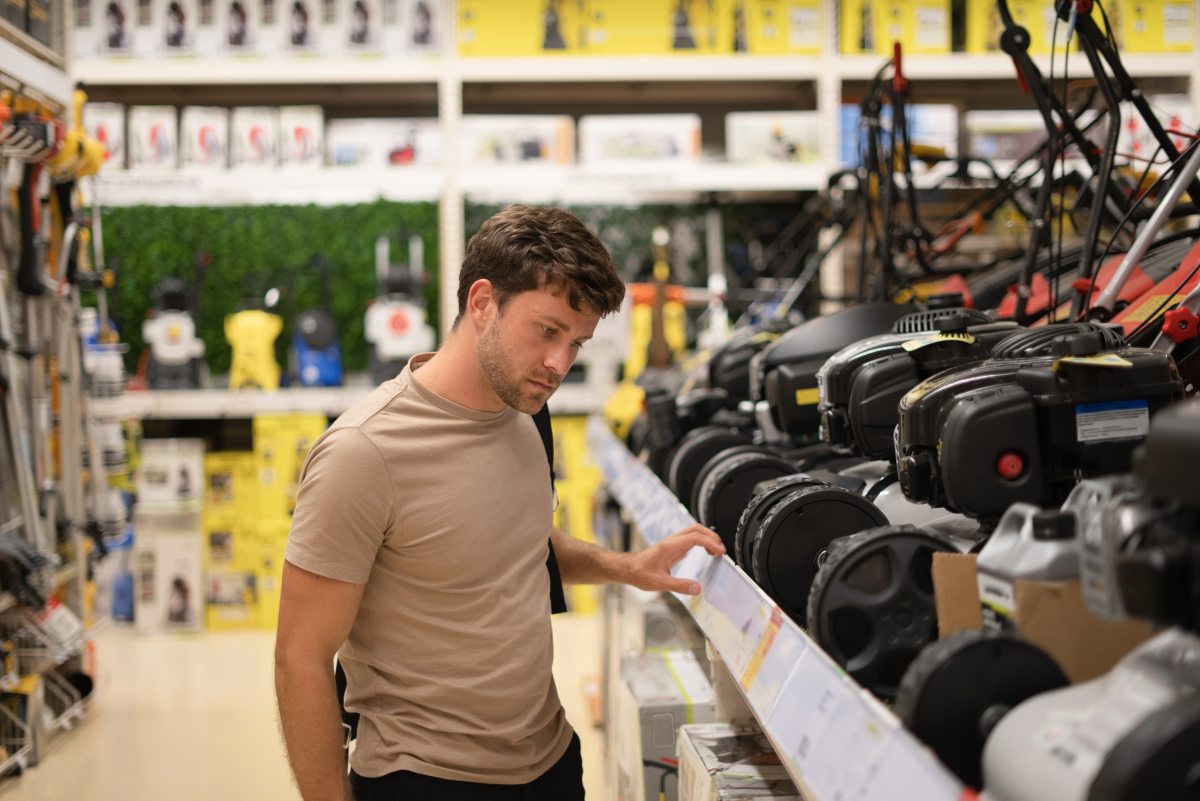 Side view of pensive casual male checking row of modern lawn mowers on shelf in power tools shop