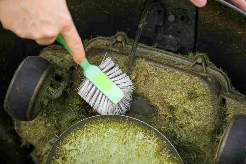 Close up view of man hand brushing off layer of wet grass stuck under automated lawnmower