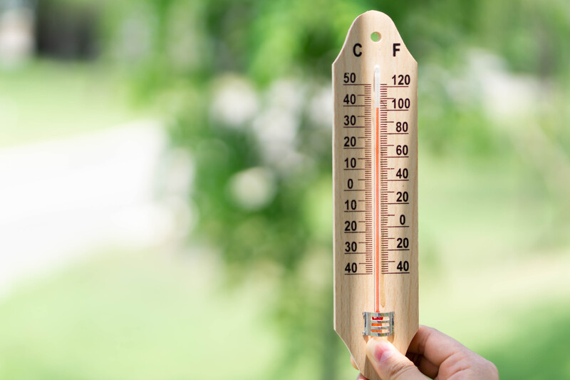 A hand holding up a wooden thermometer displaying 100 degrees Fahrenheit.